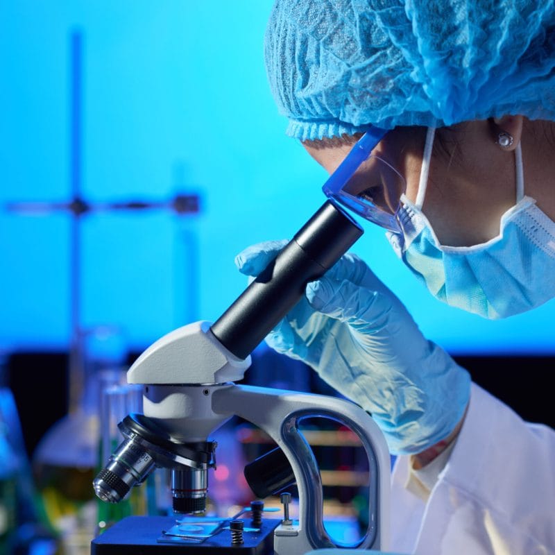 Profile view of concentrated Asian microbiologist examining sample with help of modern microscope while wrapped up in work at dim laboratory